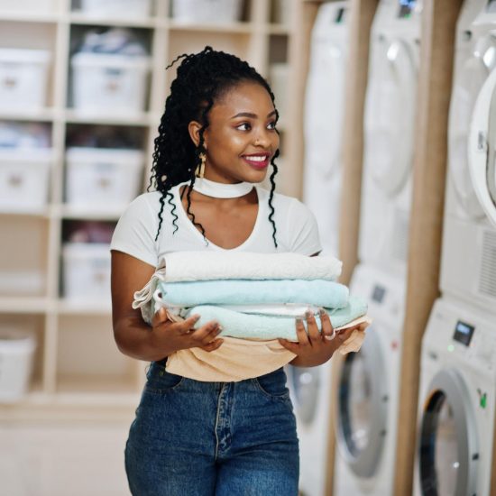 cheerful-african-american-woman-with-towels-hands-near-washing-machine-selfservice-laundry
